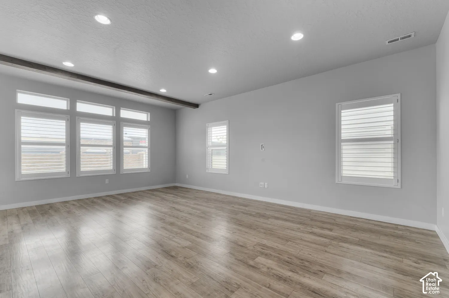 Spacious room featuring wood-finish flooring, recessed lighting, expansive windows with shutter installations, and a decorative exposed wooden ceiling beam