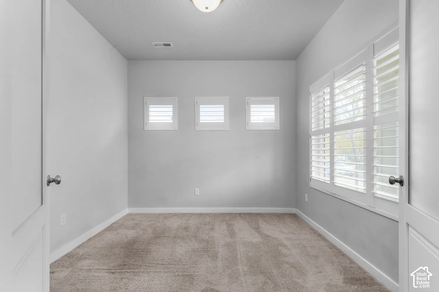 Interior room featuring wall-to-wall carpeting, three square windows with white plantation shutters, and a large multi-pane window assembly also equipped with white plantation shutters