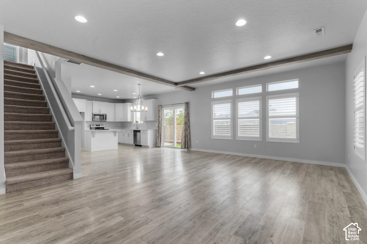 Open-concept interior featuring wood-finish flooring, recessed lighting, exposed ceiling beams, a kitchen with white cabinetry and stainless steel appliances, and a trio of windows with plantation shutters