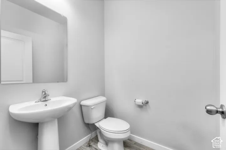 Powder room featuring a white pedestal sink with a chrome single-handle faucet, a wall-mounted mirror, a standard-height toilet, and wood-finish flooring