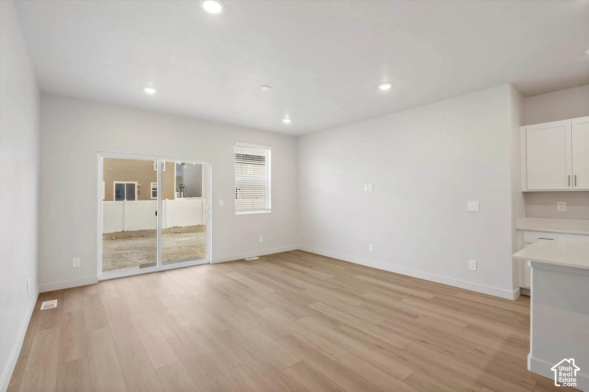 Open-concept interior featuring light wood-finish flooring, recessed lighting, sliding glass door, and white shaker-style cabinetry with solid-surface countertops