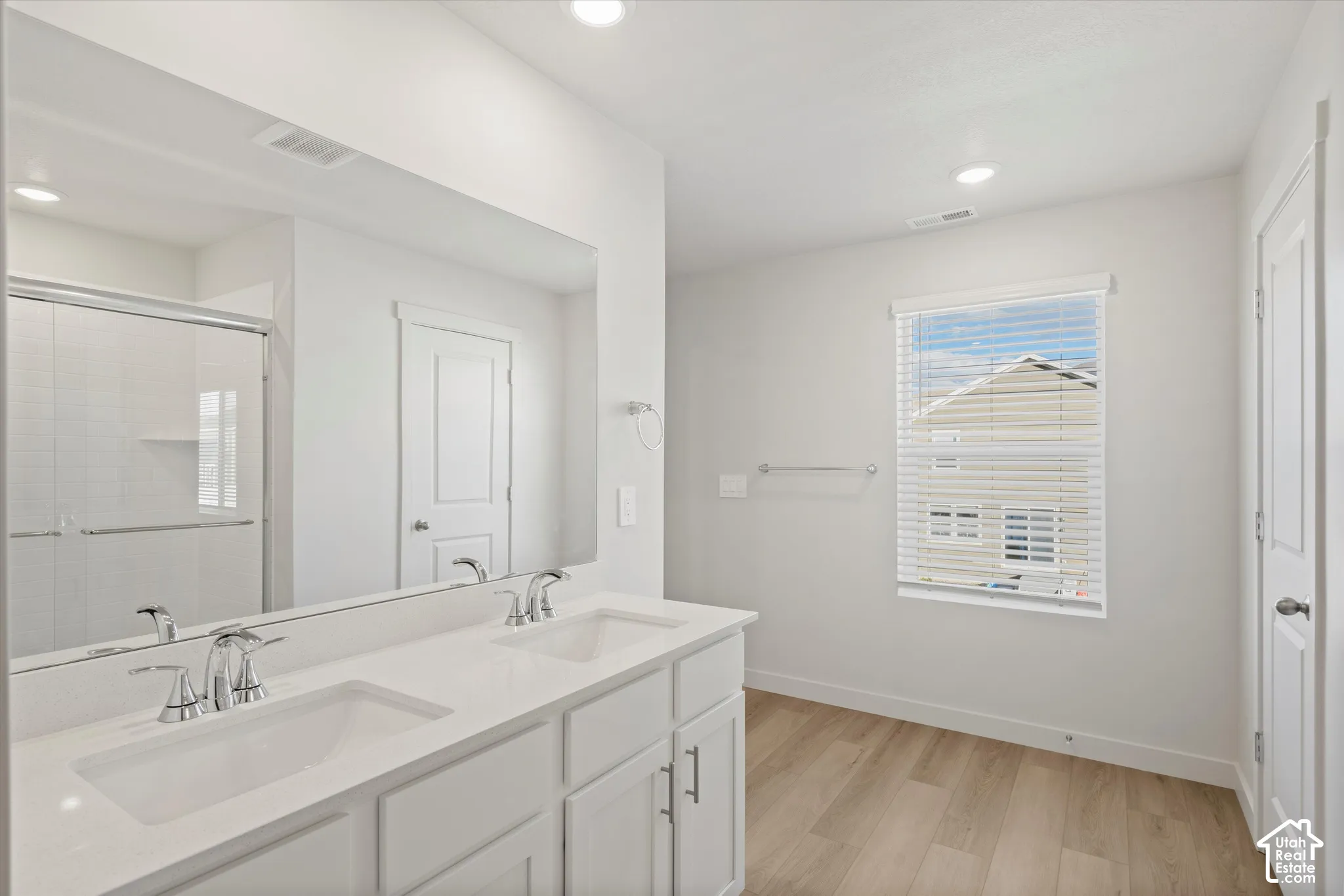Dual-sink vanity with a solid-surface countertop, full-width wall mirror, wood-finish flooring, and a glass-enclosed shower