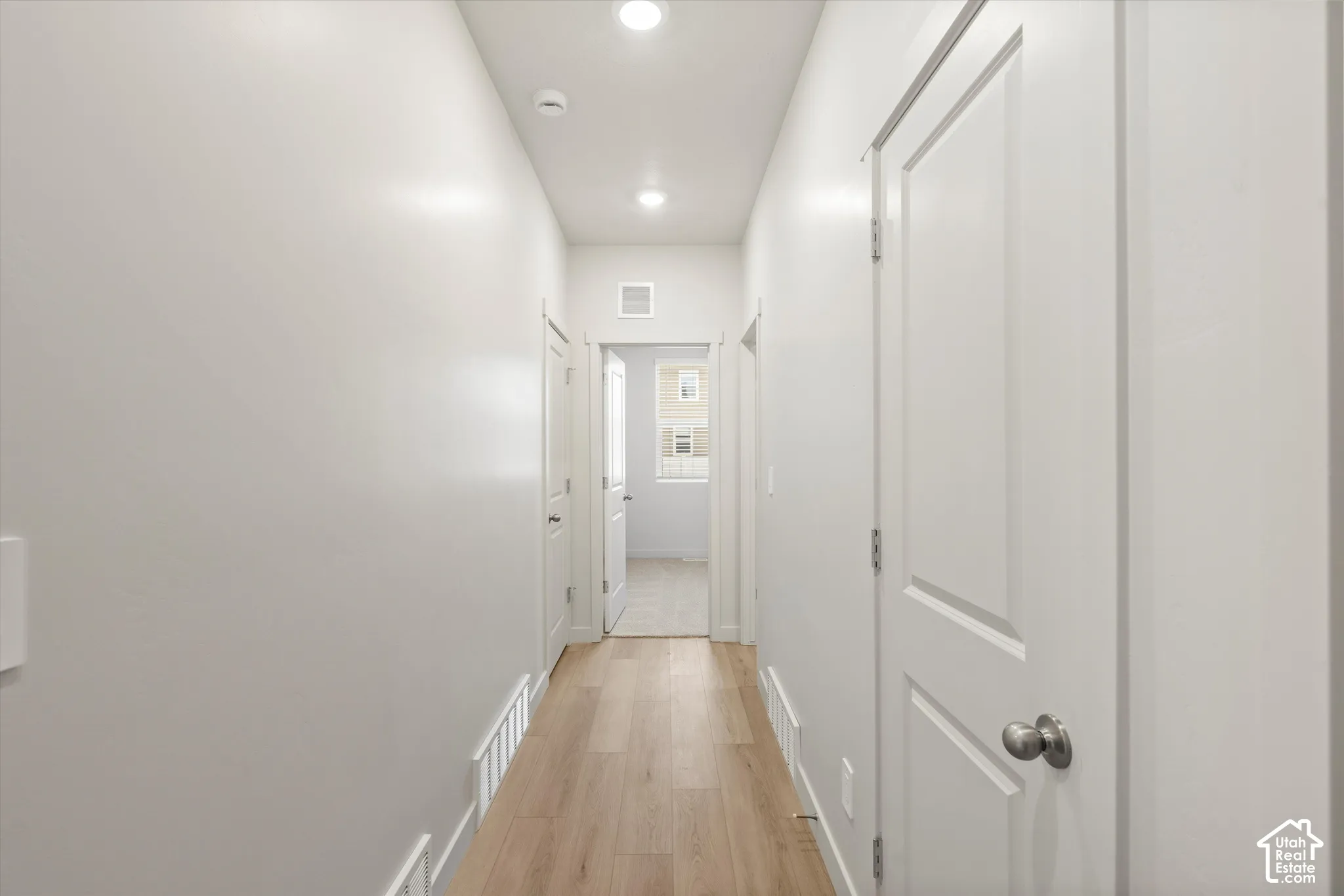 Hallway featuring light wood-finish flooring, neutral walls, recessed lighting, and white interior doors with satin nickel hardware