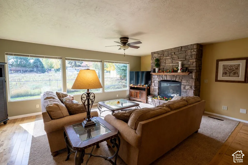 Living area featuring wood-finish flooring, a floor-to-ceiling natural stone fireplace with a wood mantel, oversized windows, and a ceiling fan