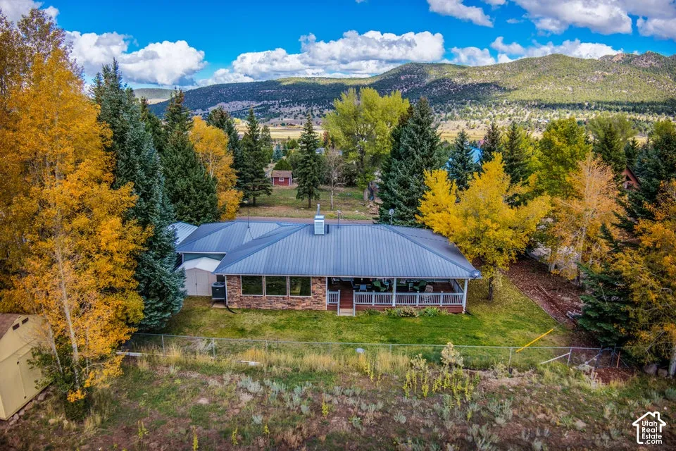 Single-story residence featuring a metal roof, stone masonry facade, covered porch with white railing, and mature deciduous and evergreen trees set against a mountain backdrop