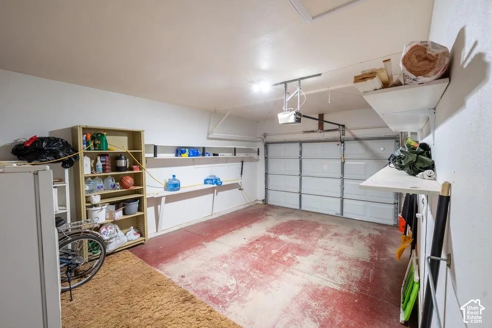 Enclosed garage with painted concrete flooring, mounted shelving, overhead door opener, and interior lighting