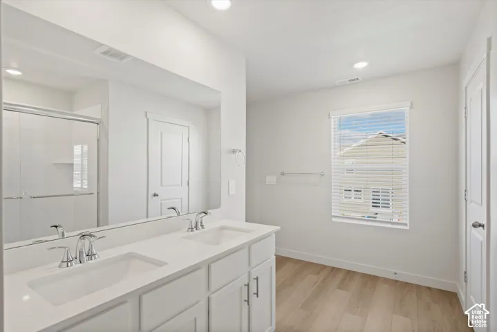 **DISCLAIMER** Interior package may vary. Dual-sink vanity with white cabinetry, light-toned countertop, and chrome fixtures, accompanied by a full-width mirror, a walk-in shower with a glass enclosure, and wood-finish flooring