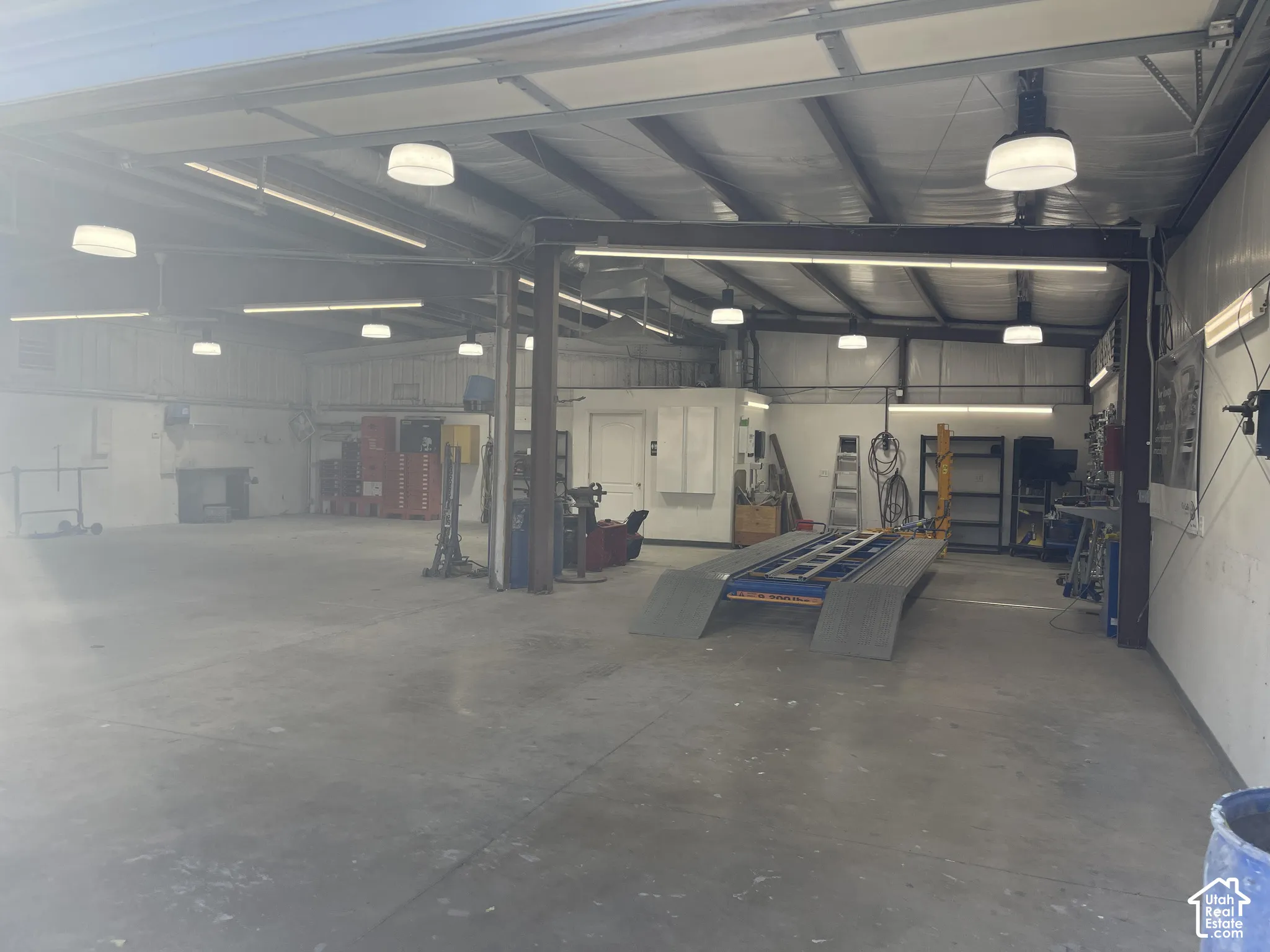 Open-span workspace featuring a concrete floor, high ceilings with exposed structural beams, industrial-style overhead lighting, and an integrated vehicle service ramp