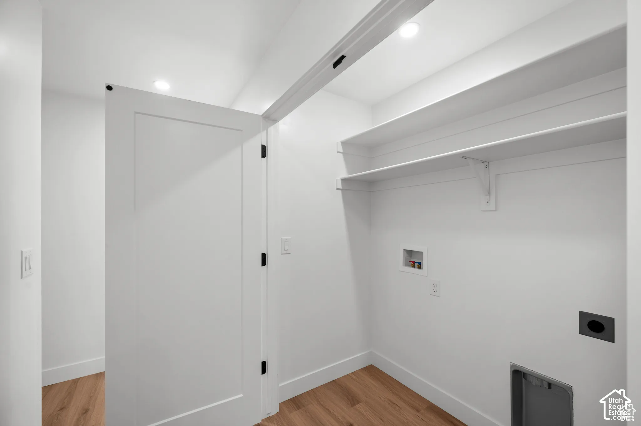 Dedicated utility space featuring wood-finish flooring, white shaker-style door, tiered open shelving, recessed lighting, and dedicated water and electrical hookups