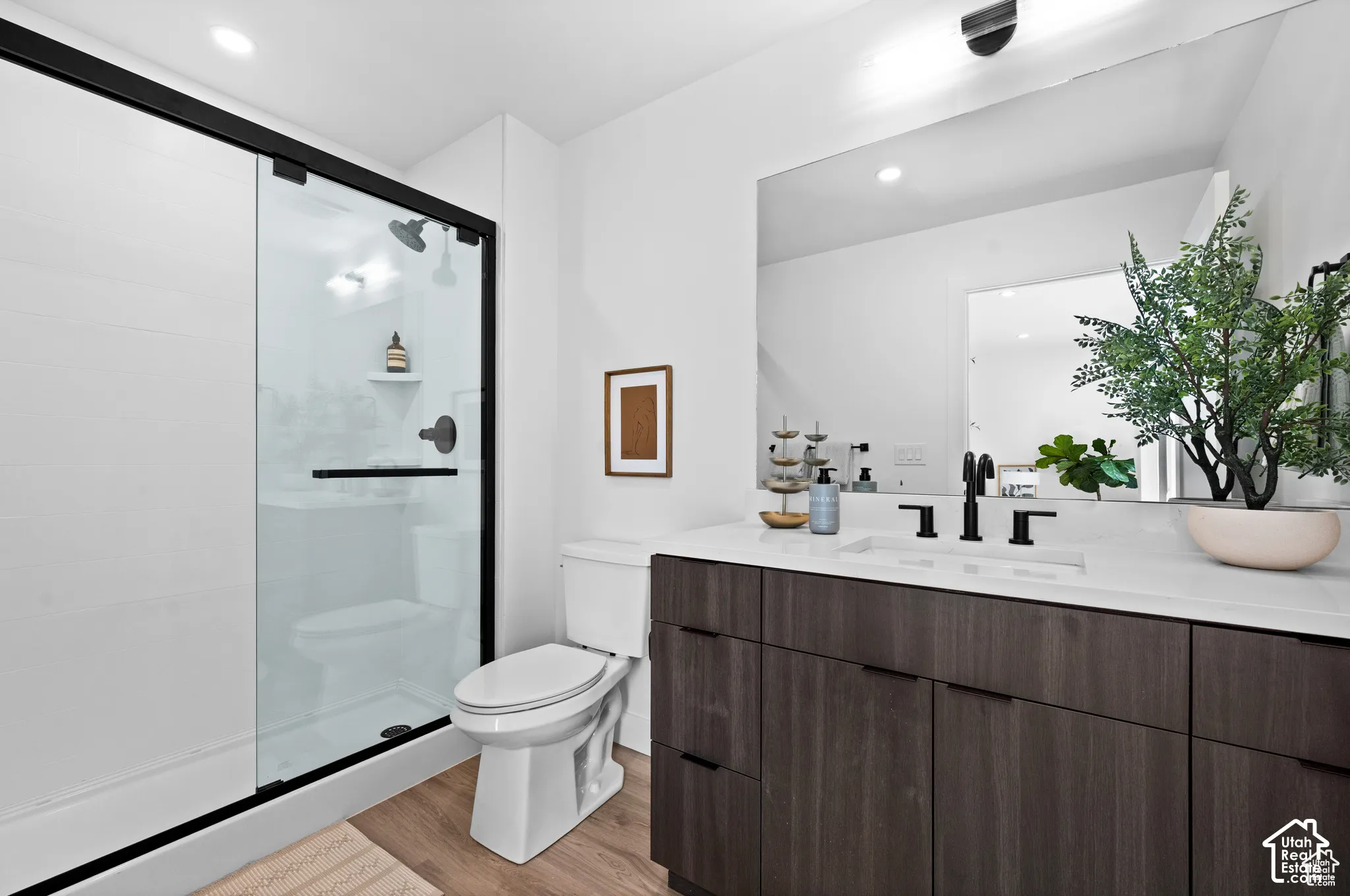 Bathroom featuring a dark wood-finish vanity with a light-toned countertop, a glass-enclosed shower with matte black hardware, a large wall-mounted mirror, and wood-finish flooring