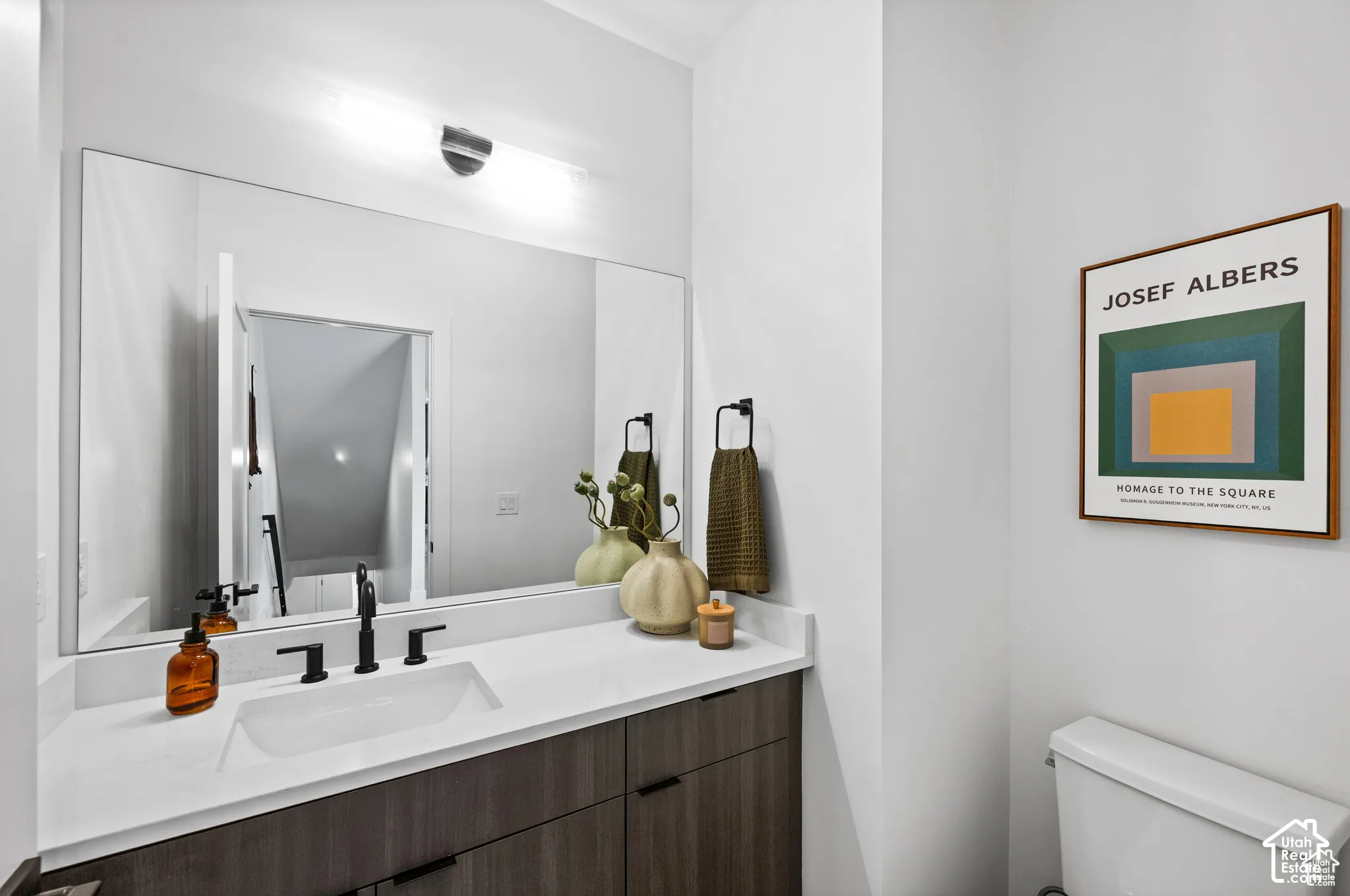 Wall-mounted lighting fixture above a large vanity mirror, dark wood-finish cabinetry, white countertop with an integrated rectangular sink, and matte black plumbing fixtures