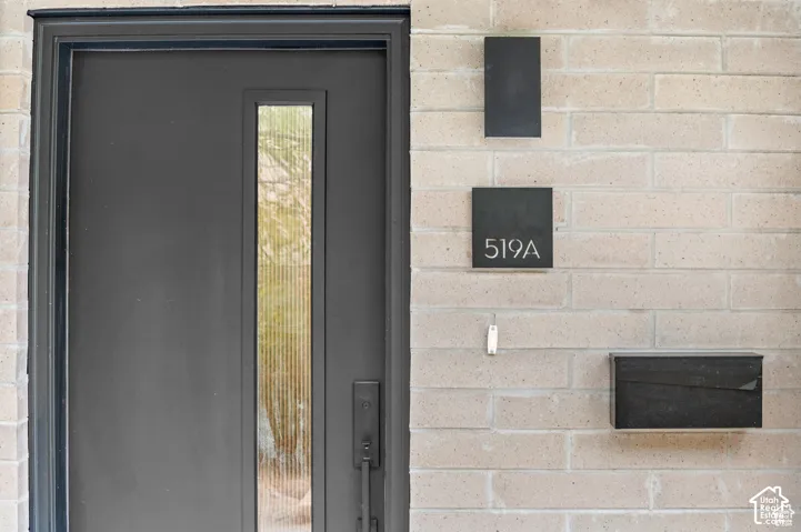 Entryway featuring a charcoal-toned door with a vertical ribbed glass panel, matte black hardware, structured exterior sconce, contemporary address plaque, and a matching wall-mounted mailbox set against a light-toned brick facade