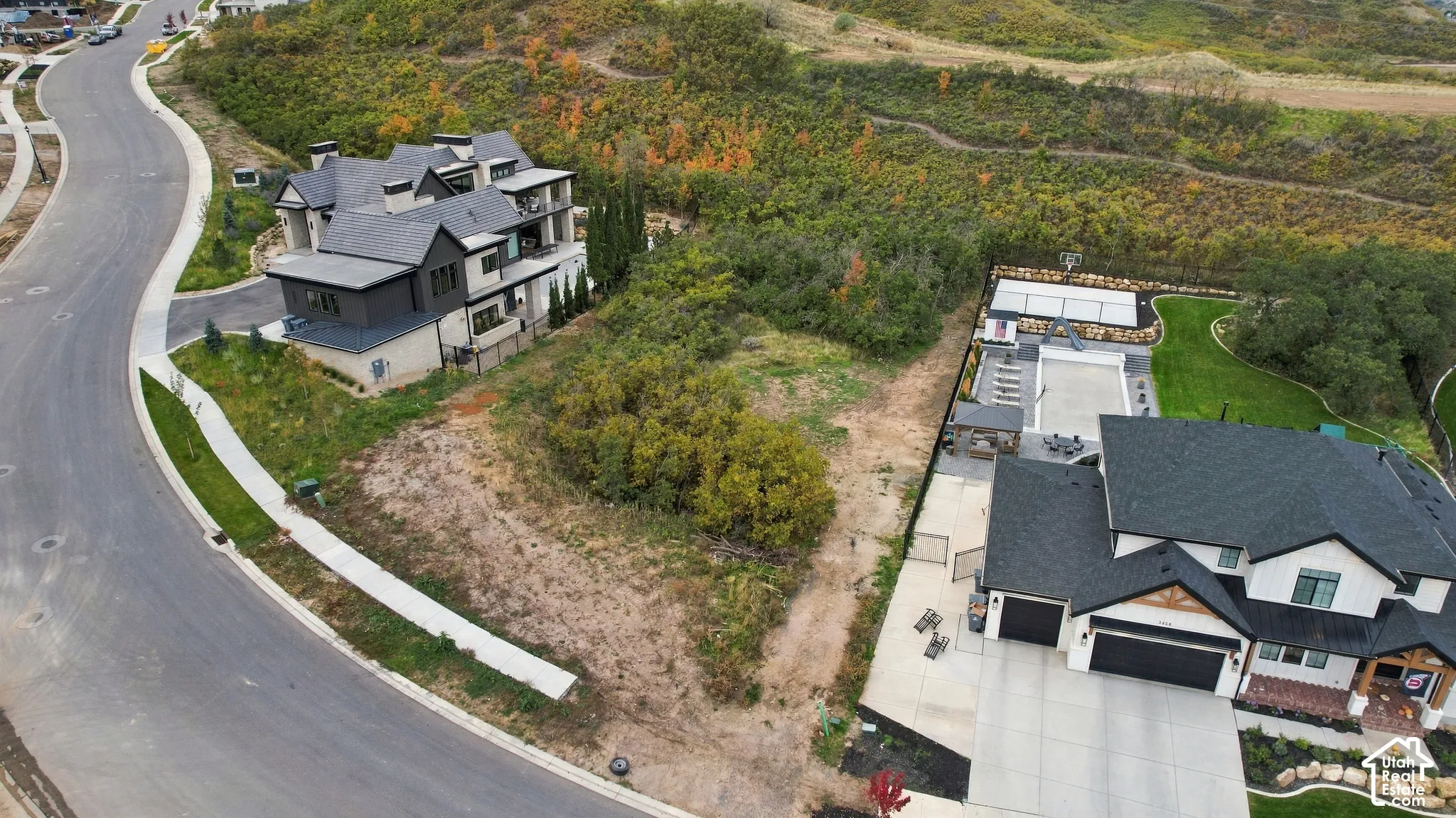 Aerial perspective of a multi-level residence featuring a multi-car garage, private backyard sport court, concrete patio, and mature hillside landscaping