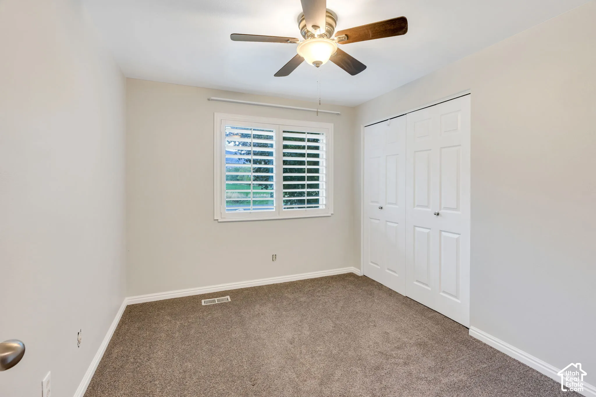 Neutral-toned bedroom featuring carpeting, a ceiling fan with light fixture, plantation shutters, and six-panel closet doors