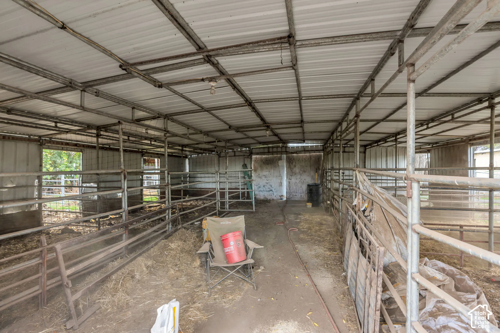 Covered livestock shelter featuring a metal roof structure, individual partitioned stalls with tubular steel fencing, and dirt flooring