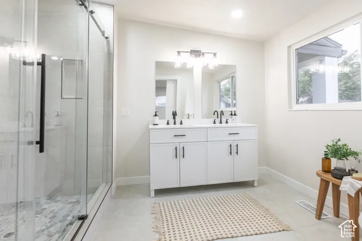 Double vanity featuring white cabinetry, stone-style countertop, and matte black hardware, paired with a glass-enclosed walk-in shower with stone tile flooring and a wall-mounted light fixture