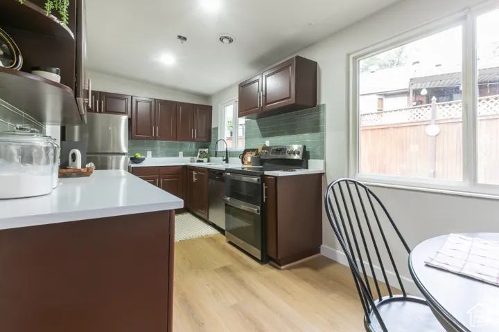 Kitchen featuring dark wood cabinetry, stainless steel appliances, light-toned solid surface countertops, a green glass tile backsplash, and wood-finish flooring