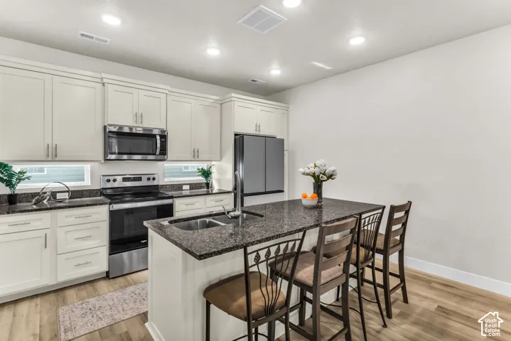 Kitchen featuring off-white cabinetry, stone-finish countertops, stainless steel appliances, a center island with an integrated sink, and wood-finish flooring