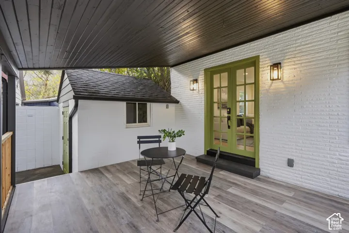 Covered patio featuring wood-finish flooring, painted brick exterior, glass-paned double doors, and integrated wall-mounted exterior lighting