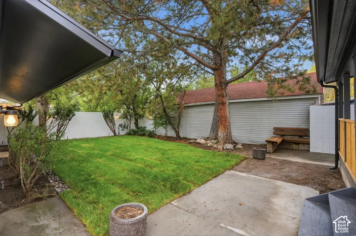Rear yard featuring a concrete patio, manicured lawn, white privacy fencing, and mature trees