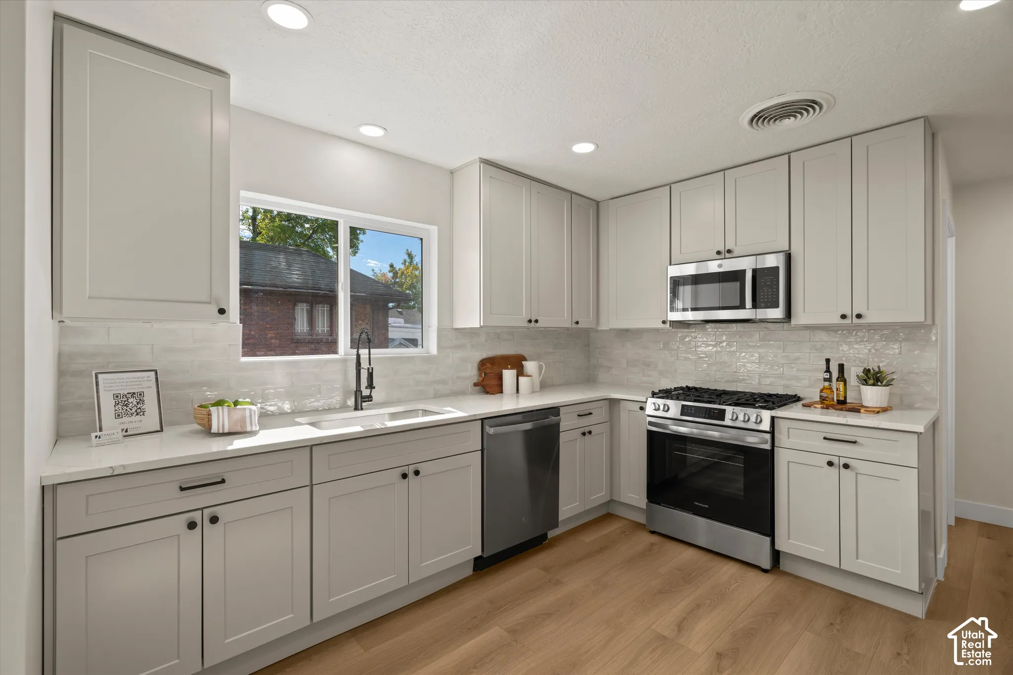Kitchen featuring light gray shaker-style cabinetry, quartz countertops, textured subway tile backsplash, stainless steel appliances, and wood-finish flooring