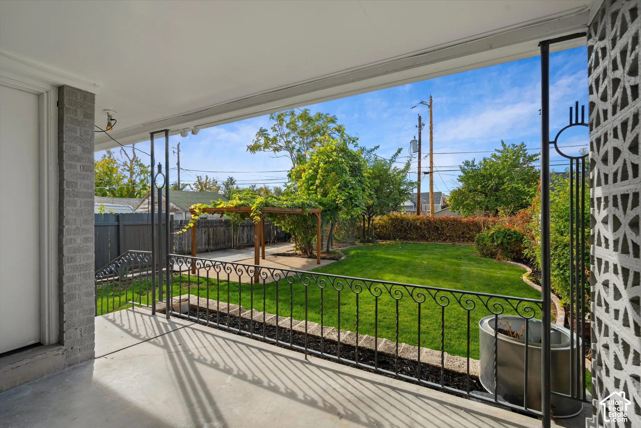 Covered concrete patio featuring decorative metal railing and patterned screen block pillars, overlooking a landscaped lawn with a wood-frame pergola and privacy fencing