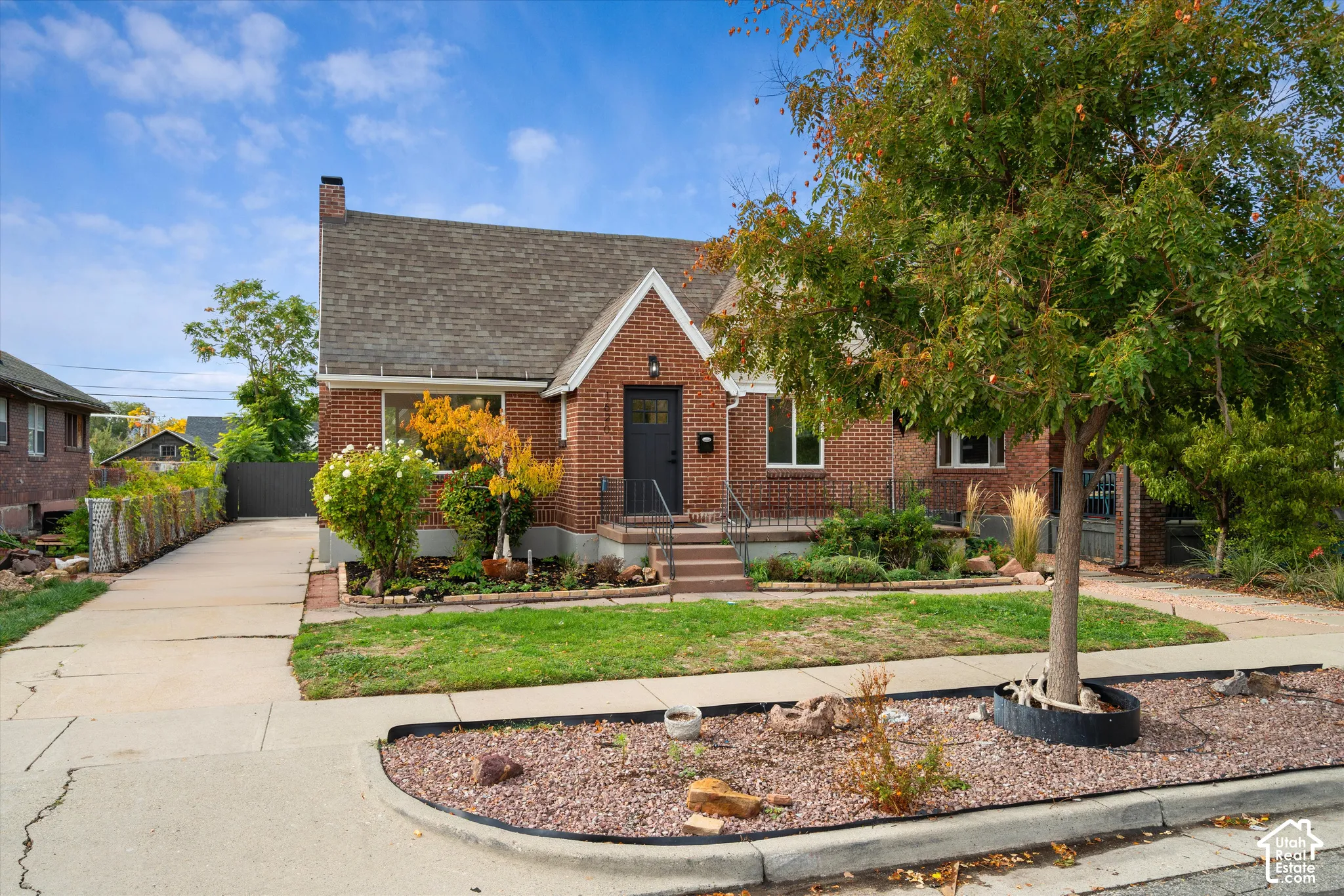 Brick exterior featuring a gabled roofline, concrete driveway, front porch with decorative metal railing, and mature landscaping