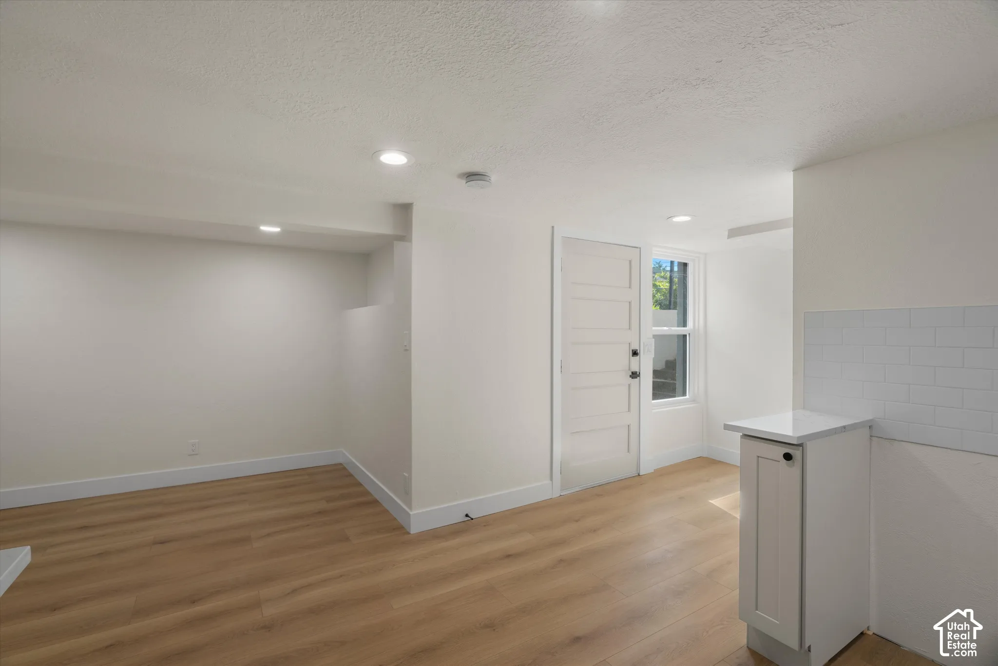 Neutral interior featuring light wood-finish flooring, recessed lighting, a single-panel door, and a kitchenette area with a white tiled backsplash