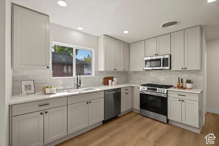 Kitchen featuring light gray shaker-style cabinetry, quartz countertops, textured subway tile backsplash, stainless steel appliances, and wood-finish flooring