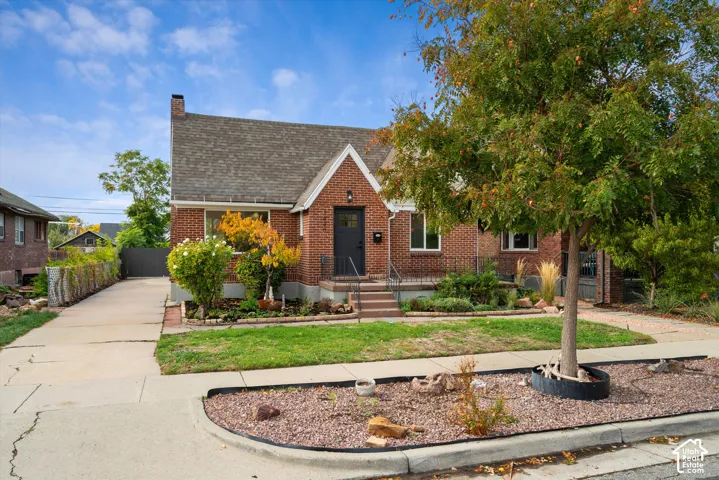Brick exterior featuring a gabled roofline, concrete driveway, front porch with decorative metal railing, and mature landscaping