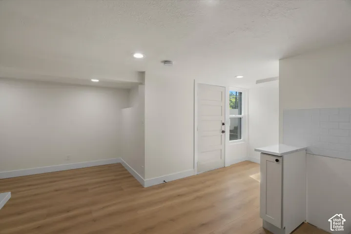 Neutral interior featuring light wood-finish flooring, recessed lighting, a single-panel door, and a kitchenette area with a white tiled backsplash