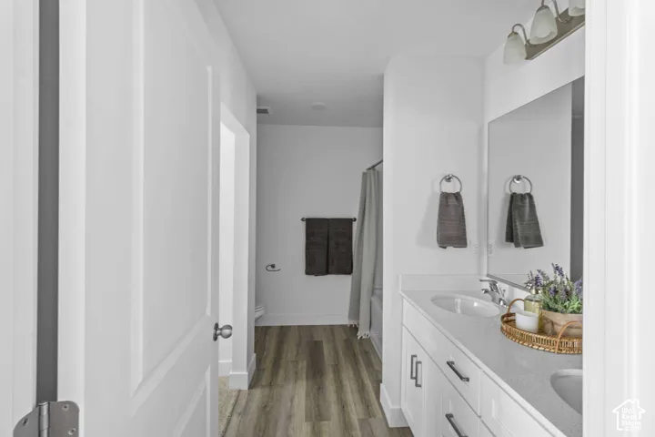 Bathroom featuring a dual-sink vanity with light-toned solid surface countertops, white shaker-style cabinetry with matte black hardware, wood-finish flooring, and a large wall-mounted vanity mirror with a three-light fixture