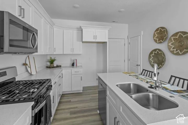 Kitchen featuring white cabinetry, quartz countertops, stainless steel appliances, a center island with a dual-basin sink, and wood-finish flooring