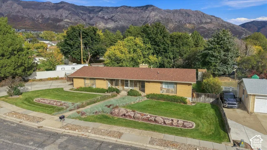 Single-level brick exterior featuring a shingle roof, mature landscaping, stone garden beds, and a detached garage, set against a mountain backdrop