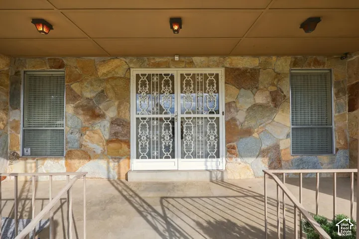 Exterior entryway featuring a natural stone facade, double front doors with decorative metal security grilles, two windows with horizontal blinds, and a covered porch with metal railings