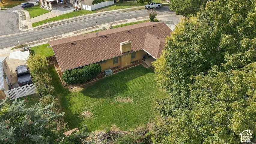 Aerial perspective of the property featuring a structured roofline with a central chimney, concrete driveway, white vinyl fencing, and a manicured lawn framed by mature trees