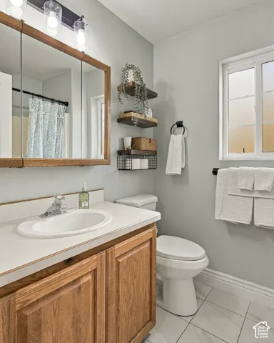 Bathroom featuring wood-finish cabinetry, a white countertop with an integrated oval basin, light-toned tile flooring, three-light vanity fixture, and tiered wall-mounted shelving