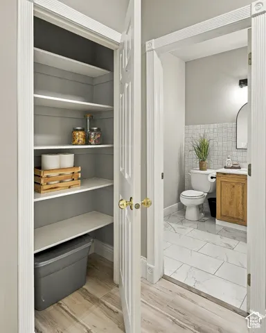 Storage closet with built-in shelving featuring white-finish planks, two-panel interior doors with brass hardware, bathroom vanity with wood-finish cabinetry, white stone-pattern tile flooring, and decorative grid-pattern wall tile
