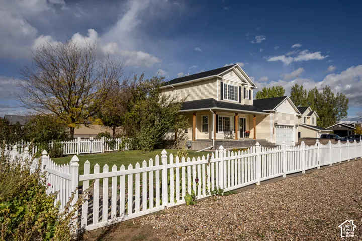 Two-story residence featuring light-toned horizontal siding, a covered front porch with wood-finish structural columns, a white vinyl picket fence, and a multi-car garage