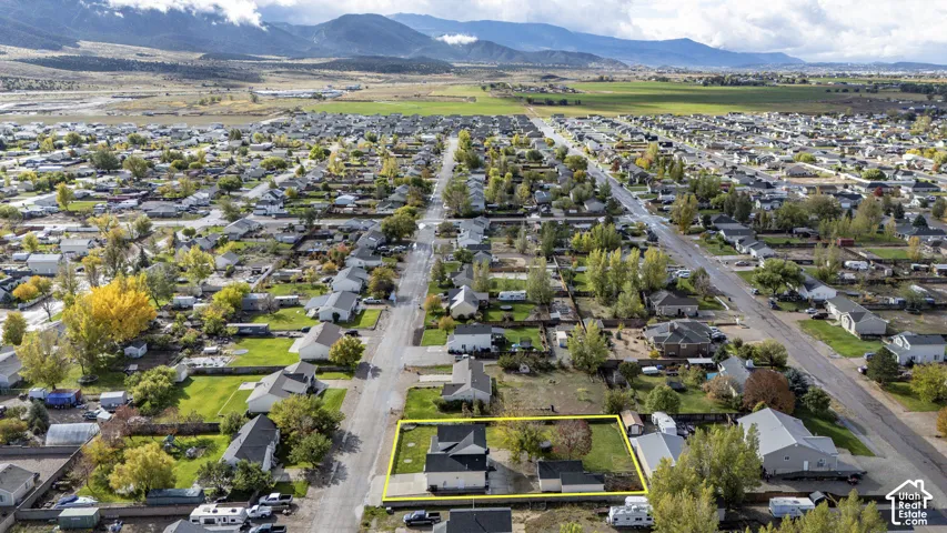 Aerial perspective featuring a residential lot with a paved driveway, detached structure, and open green space, set against a mountainous horizon