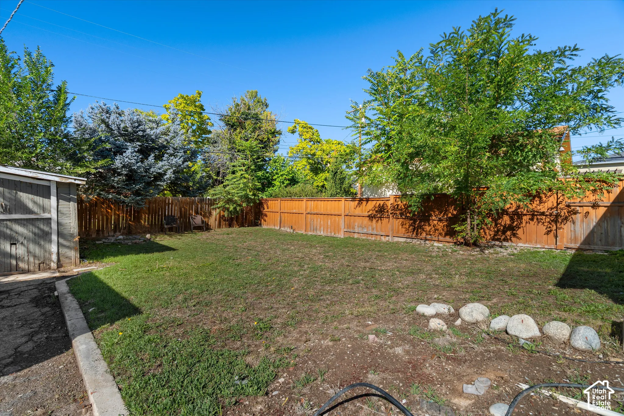 Backyard featuring a grass lawn, perimeter wood fencing, mature trees, and stone border detailing