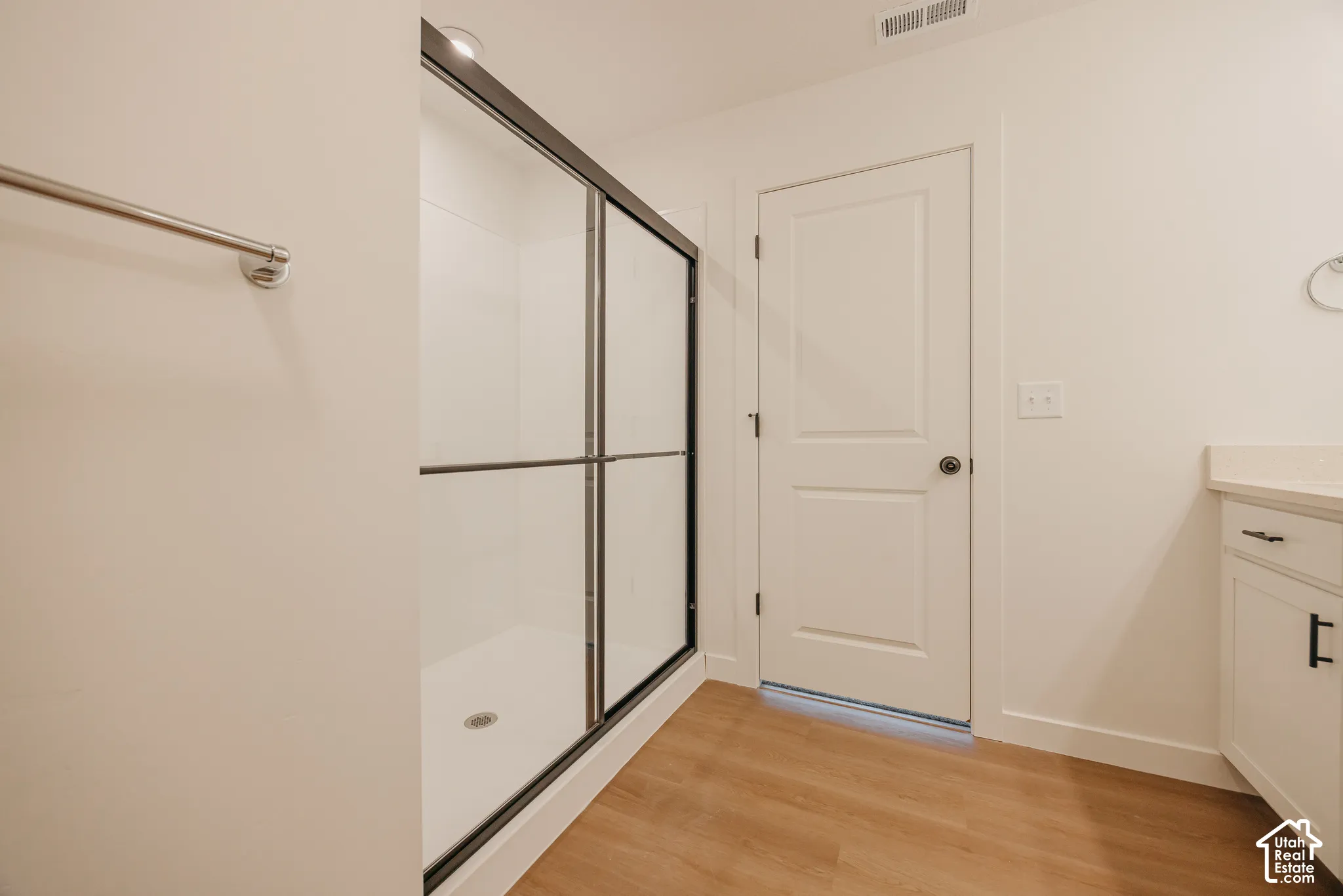 Shower enclosure with a black frame and sliding glass doors, white cabinetry with matte black hardware, light-toned wood-finish flooring, and a solid-panel door