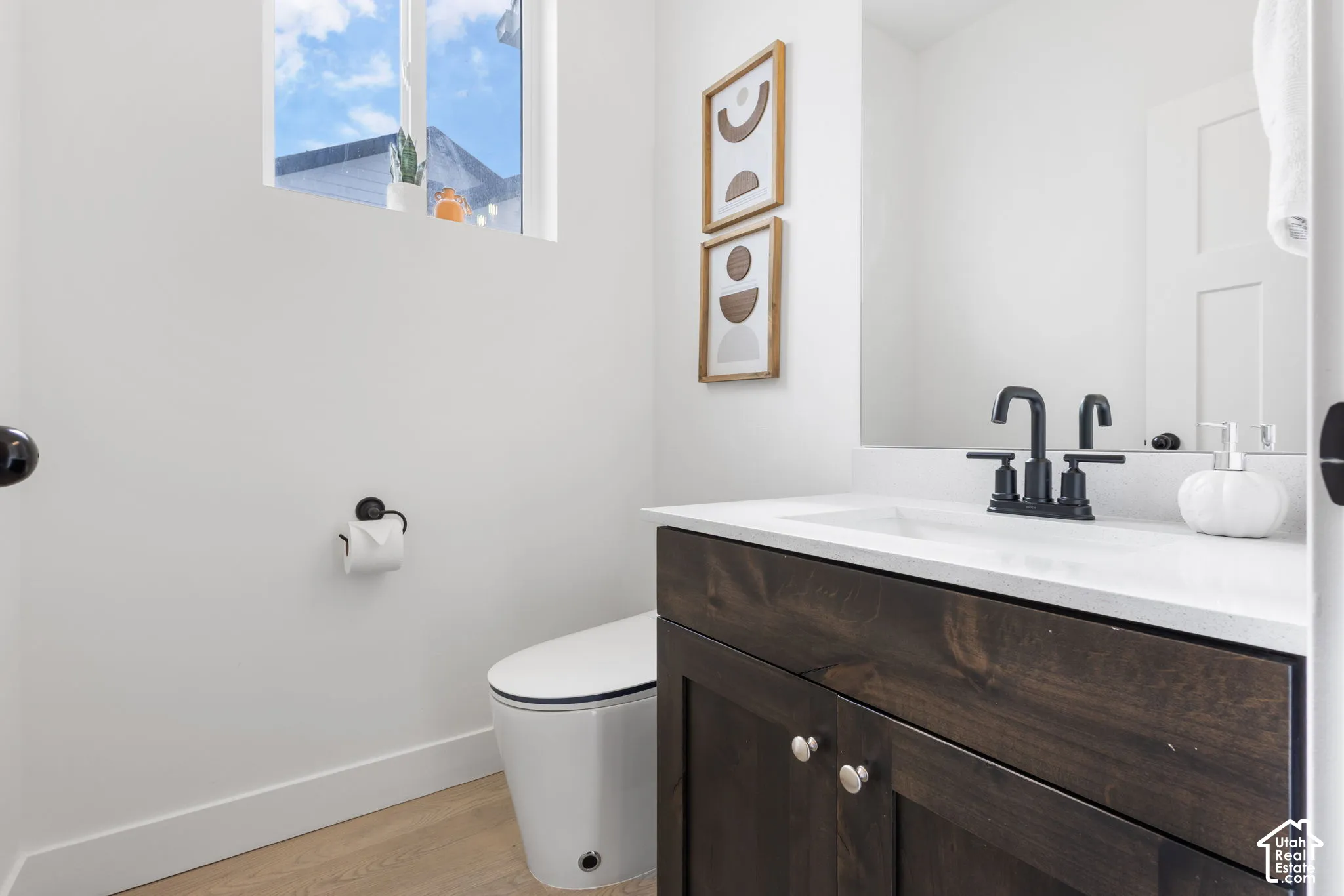 Dark wood-finish vanity with a white countertop and rectangular undermount sink, accented by a matte black widespread faucet, large wall mirror, and light-toned wood-finish flooring