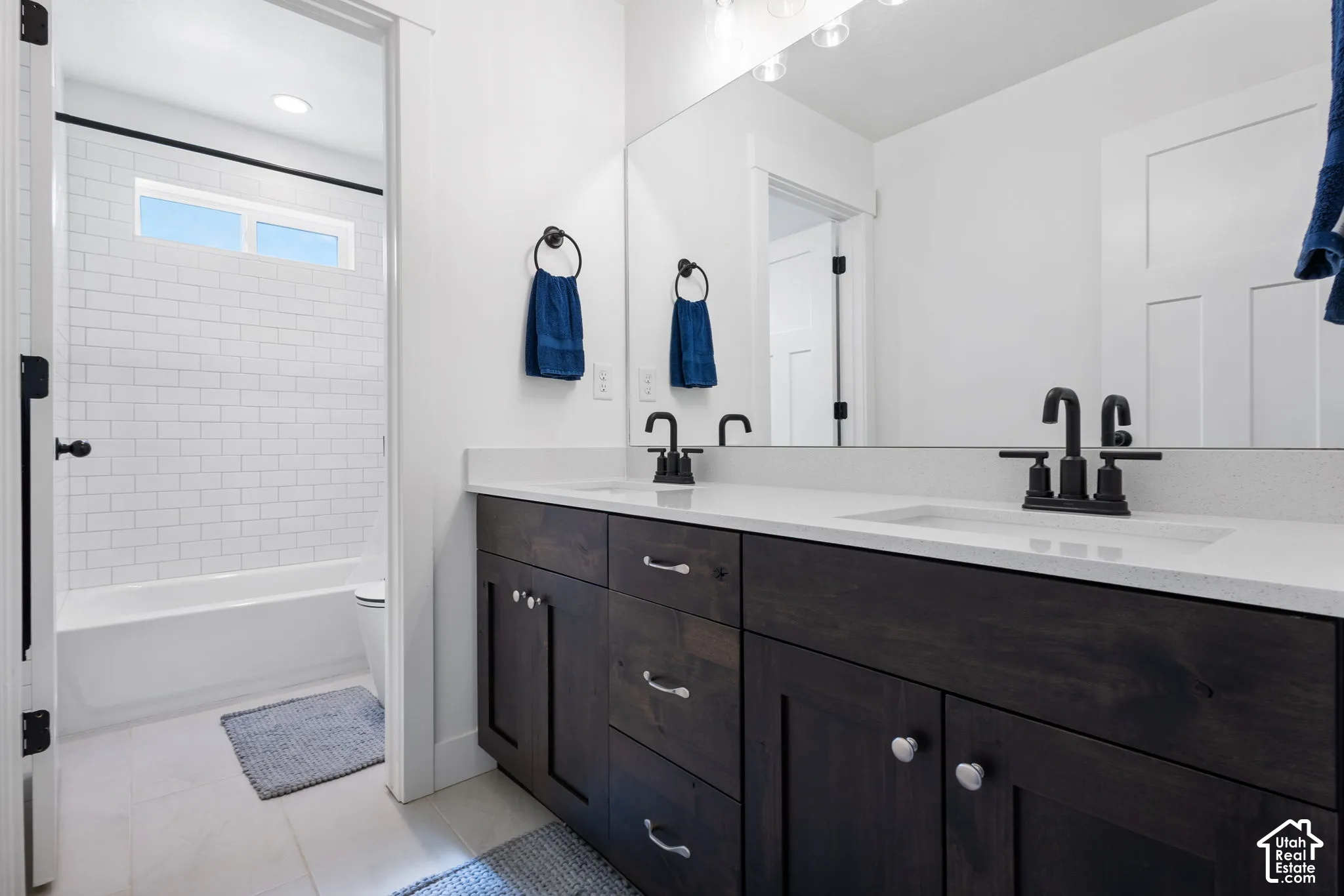Dual-sink vanity with dark-finish cabinetry, light stone-composite countertop, matte black fixtures, and full-width vanity mirror