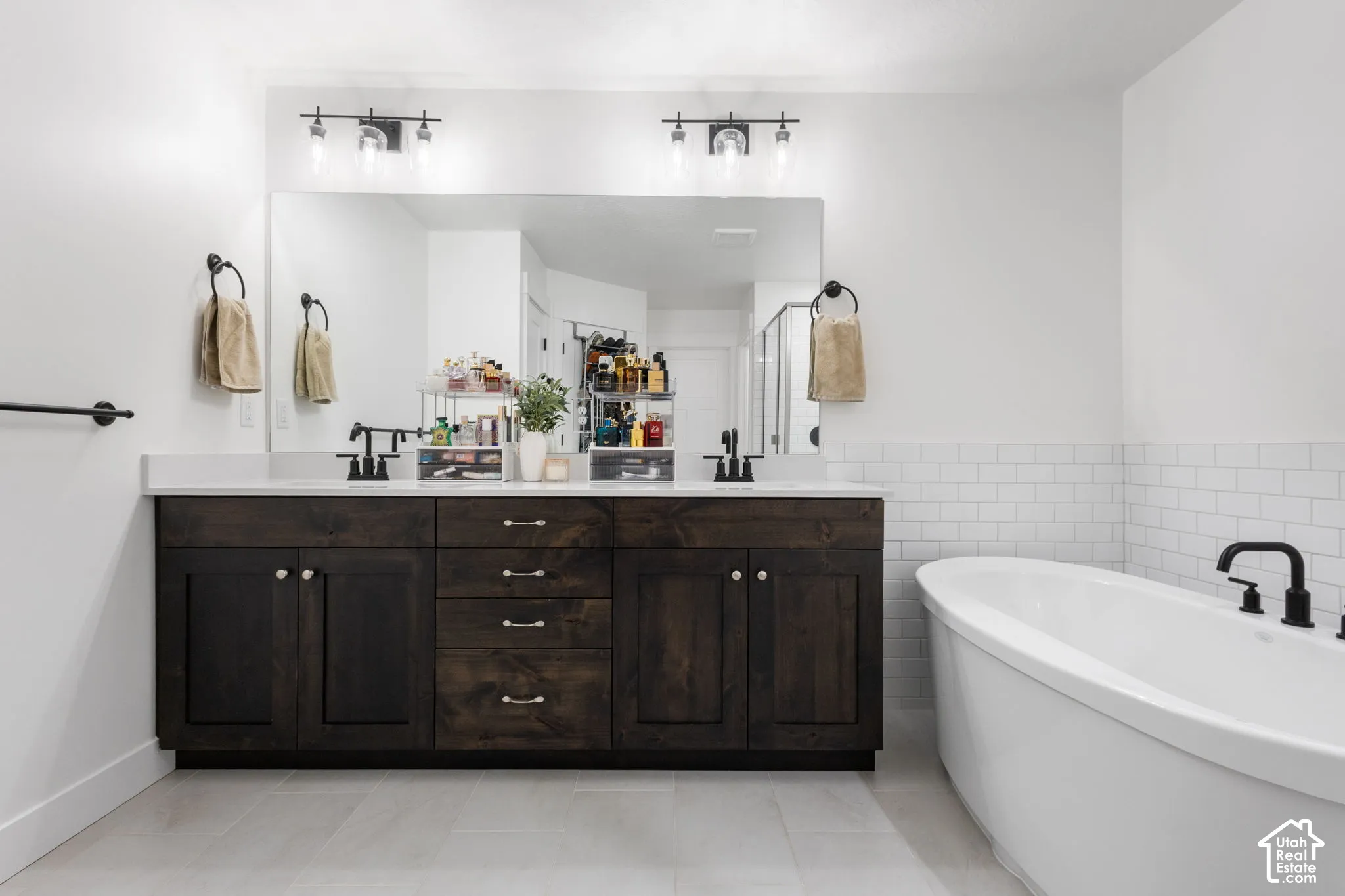 Double vanity with dark wood-finish cabinetry, solid surface countertop, matte black fixtures, and subway tile backsplash featuring a deep soaking tub