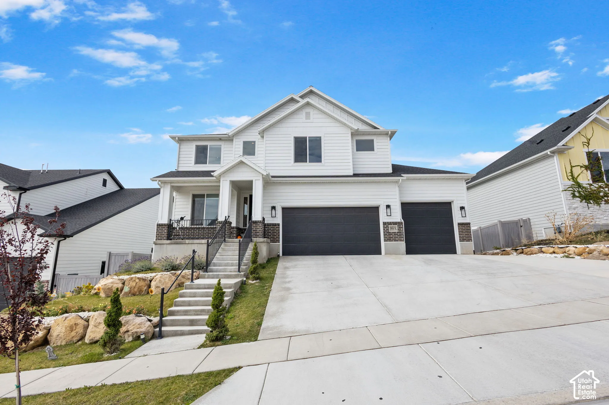 Two-story residence featuring white horizontal siding, dark brick veneer accents, a three-car garage, and concrete hardscaping with stone retaining walls