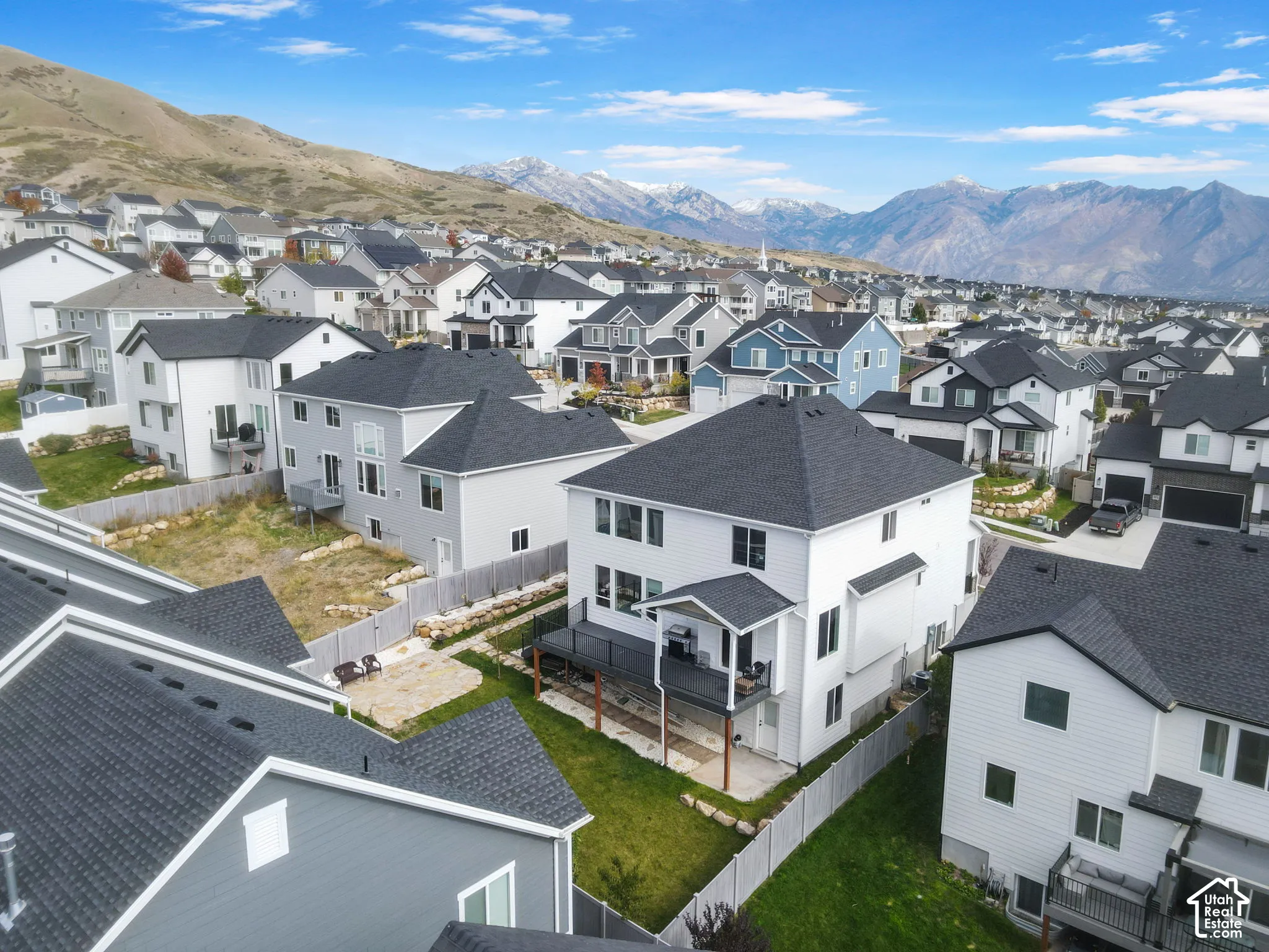 Aerial perspective of a multi-story residence featuring a raised back deck, ground-level patio, vinyl siding, shingle roof, and a fenced backyard against a mountain backdrop