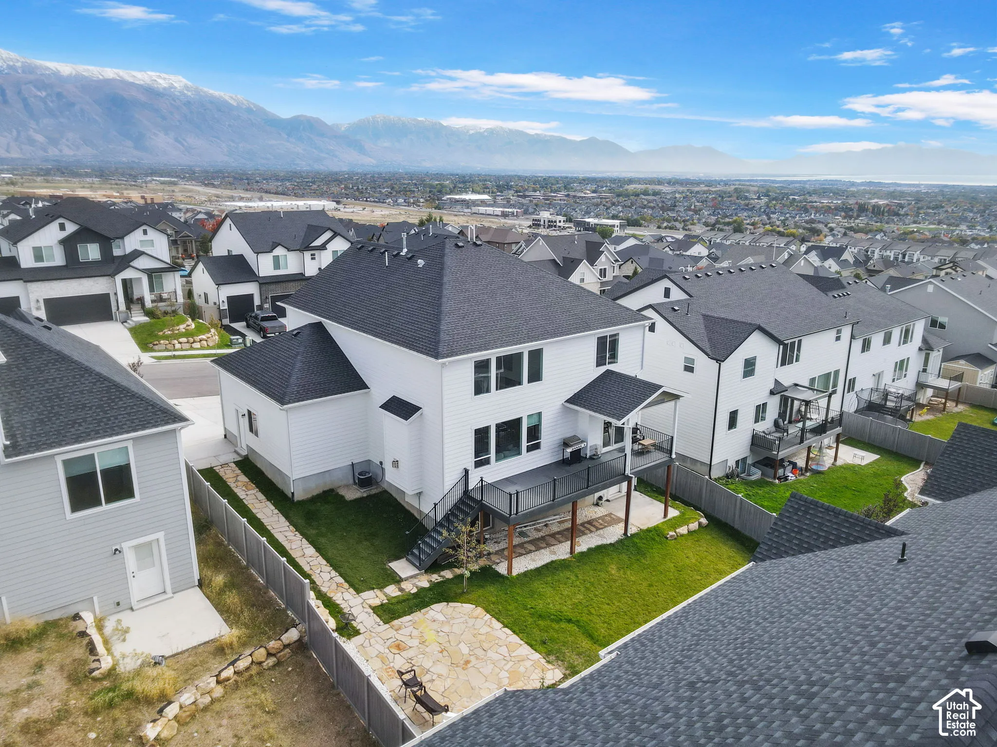 Two-story residence featuring white siding, a dark shingle roof, an elevated composite deck with metal railings, a stone paver patio, and a grass lawn set against a mountain backdrop
