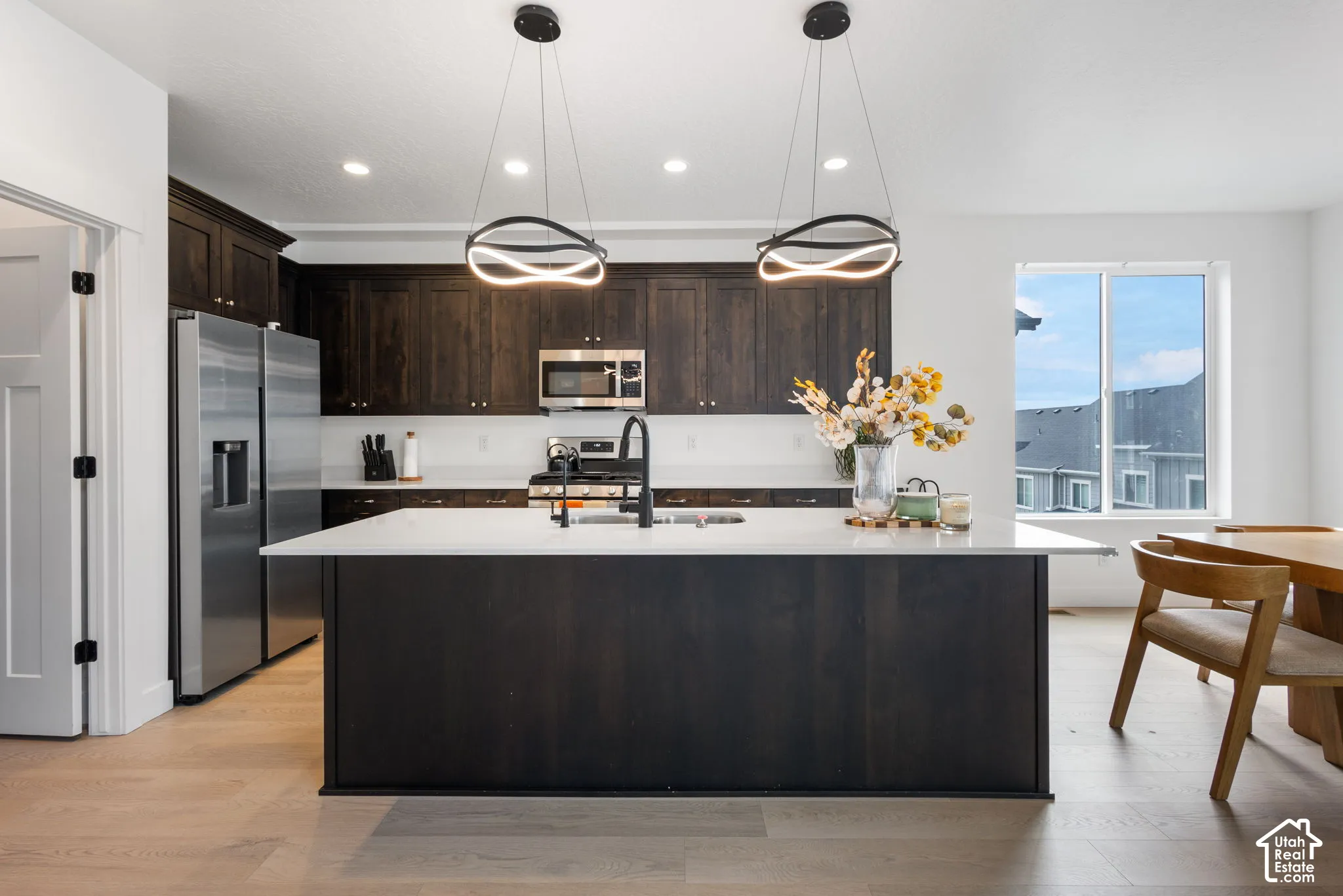 Kitchen featuring dark-stained cabinetry, a large central island with a white solid-surface countertop, stainless steel appliances, modern circular pendant lighting, and light-toned wood-finish flooring
