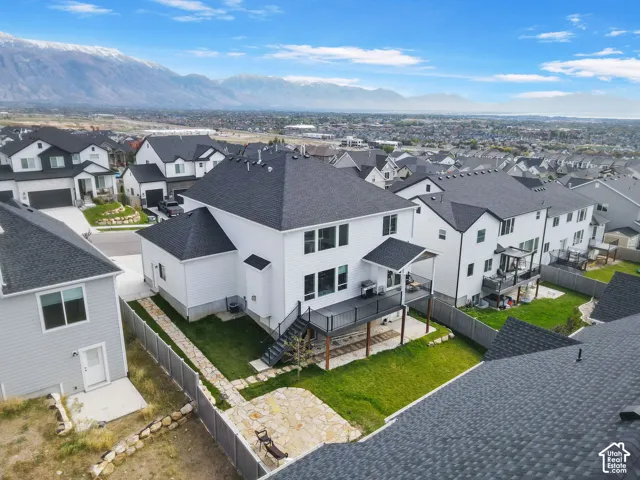 Two-story residence featuring white siding, a dark shingle roof, an elevated composite deck with metal railings, a stone paver patio, and a grass lawn set against a mountain backdrop